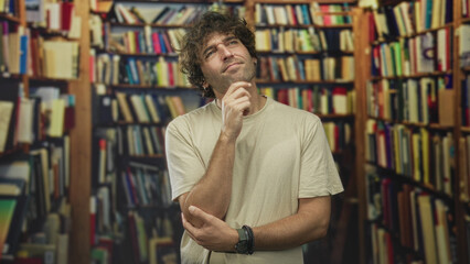 Young hispanic man touches chin in a library building surrounded by bookshelves and books  thoughtful curiosity. © Krakenimages.com