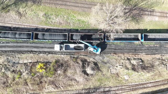Aerial view of dump truck being loaded with coal from freight train cars using a crane, showcasing the continuous action of material transfer in an industrial setting