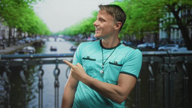 Man referee in turquoise shirt points finger and displays whistle on lanyard on a street bridge by a canal, smiling and speaking; friendly greeting.