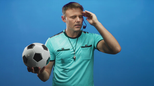 Man holding a soccer ball in one hand and touching his temple with the other hand, referee with whistle and headset visible in studio; focus strategy.