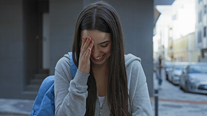 Fototapeta premium Young woman with backpack laughing and covering her face with one hand on a city street; youthful happiness.