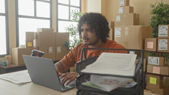 Man in orange shirt holding document and hand on cheek beside laptop and barcode scanner in building; small business concentration.