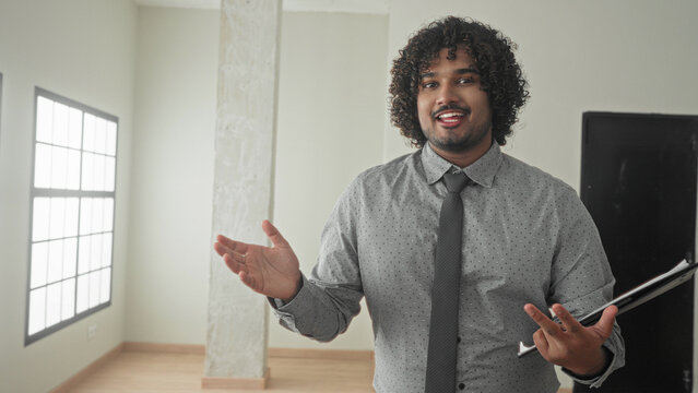 Young man holding clipboard and pointing finger in empty building interior with large windows and wooden floor; trust presentation confidence.