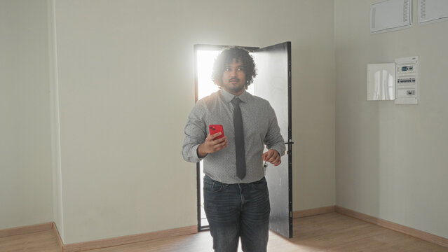 Man holding smartphone and keys, hand extended while entering a vacant building with open door and sunlight behind him; new home optimism.