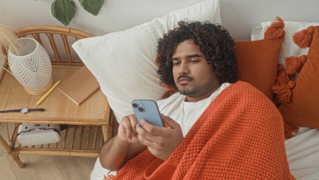 Man indian young lying in bed under an orange blanket holding smartphone and pill bottle, reading label by bedside table in a building; concern selfcare.