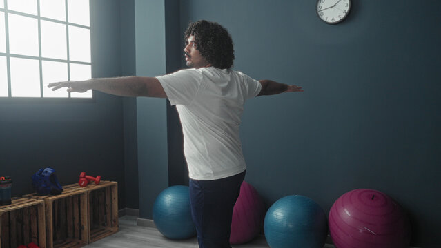 Man in white t-shirt, arms outstretched, balancing near blue and pink exercise balls and red dumbbells on wooden crates beside a large window and clock in a studio; calm focus balance.