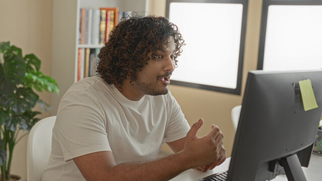 Young indian man with hands over mouth, seated at a desktop computer reacting to bright office in a building with bookshelf and plant visible; surprise success.