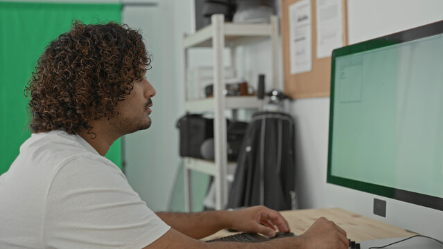 Man photographer seated at desk types on keyboard and turns editing dial with hands beside dslr camera and large monitor against green screen and shelves in studio; concentration.