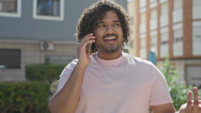 Man holding smartphone to ear, smiling and gesturing on a sunny street in front of buildings; friendly conversation cheerful.
