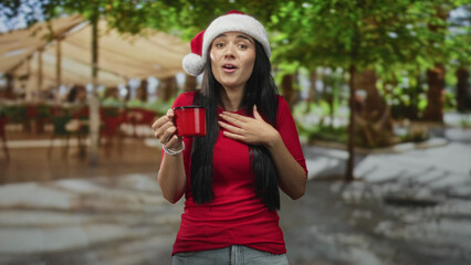 Naklejka premium Young beautiful hispanic woman in santa hat holds red mug, hand to chest and eyes closed in a street cafe with canopy and trees; holiday joy.