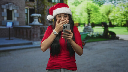 Naklejka premium Woman in santa hat and red top taps smartphone with fingers while smiling in a building courtyard near steps and fountain; holiday joy.