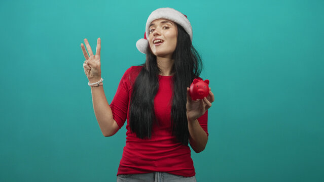 Young woman wearing santa hat holds red piggy bank and shows peace sign in studio; festive savings cheer.