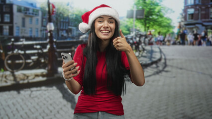 Naklejka premium Woman wearing santa hat holding smartphone and giving thumbs up on cobblestone street by a canal with bicycles; festive joy.