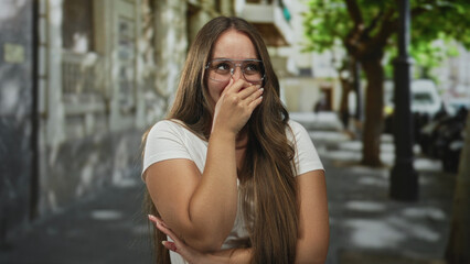 Fototapeta premium Woman covers mouth with hand on street wearing glasses and white t shirt; playful embarrassment shyness.