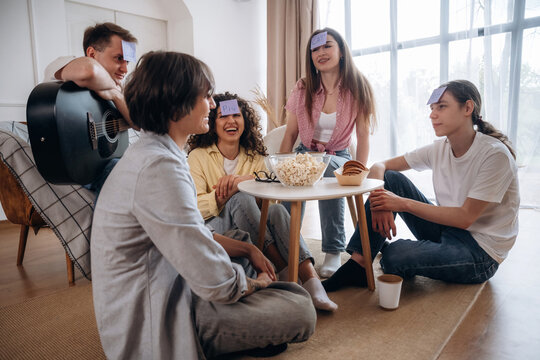 Group of young adults playing a guessing game with sticky notes on their foreheads, sitting on the floor in a cozy living room with natural light and snacks on the table