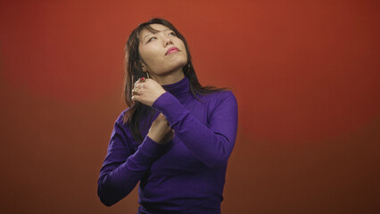 Fototapeta premium Woman flips hair with hand and tilts head wearing purple turtleneck and hoop earring against red studio backdrop; confidence poise.