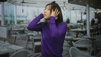 Fototapeta premium Woman covering ears with hands and looking upward inside a bright restaurant building; anxiety overwhelm.