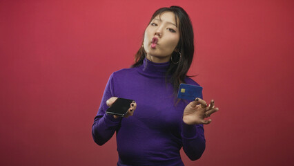 Woman holding smartphone and creditcard while puckering lips in studio with red backdrop and hoop earrings  playful confidence. © Krakenimages.com