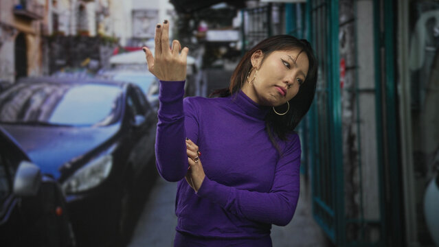 Woman touching her arm with hand and rings beside parked car on a narrow street, wearing purple turtleneck; contemplation.