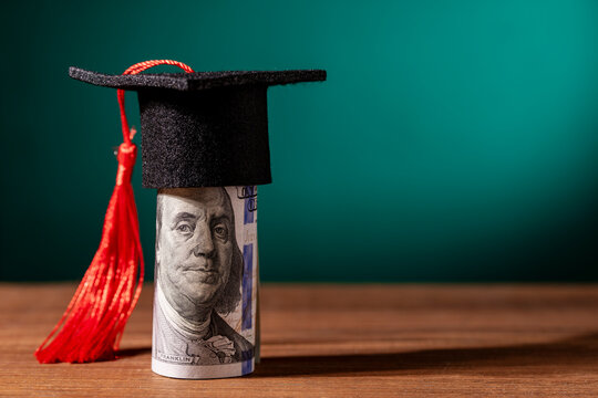 Graduation cap on hundred dollar bill. Miniature graduation mortarboard hat on rolled 100 dollar banknote. Concept of high cost of education student loans university tuition and financial success