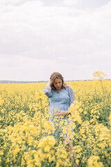 Fototapeta premium A beautiful pregnant woman with long wavy hair, wearing a light blue floral dress, gently holding her belly while standing in a blooming yellow rapeseed field. 