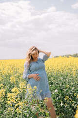 Fototapeta premium A beautiful pregnant woman with long wavy hair, wearing a light blue floral dress, gently holding her belly while standing in a blooming yellow rapeseed field. 