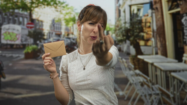 Woman in white blouse holding brown envelope shows middle finger on street outside cafe building; defiance rebellion.