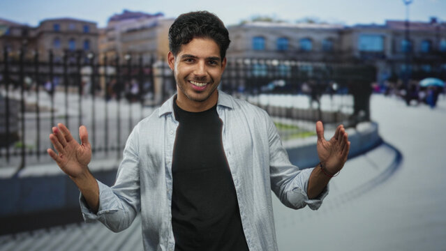 Young hispanic man smiling and showing open hands with palms outward, wearing a black tee and unbuttoned light shirt in front of an iron fence on a street; confidence.