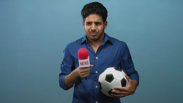 Man holding red microphone and soccer ball in studio while looking down and gripping ball with both hands; nervous interview practice.