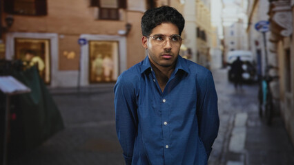 Obraz premium Young man with glasses standing with hands behind back on a narrow european street with shopwindows and cobblestones; pensive solitude.