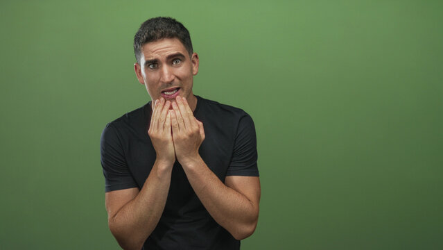 Young hispanic man with hands covering mouth and tense expression in green studio; nervousness uncertainty.