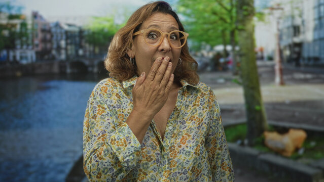 Hispanic middle aged woman covers mouth with hand on a canal street in amsterdam; unexpected astonishment.