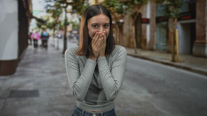 Fototapeta premium Woman covering mouth with hand in a surprised gesture on a city street near buildings; surprise privacy vulnerability.