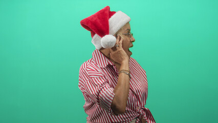 Naklejka premium Woman senior hispanic wearing santa hat cups ear with hand and open mouth, red striped shirt tied at waist against green studio backdrop; holiday surprise.
