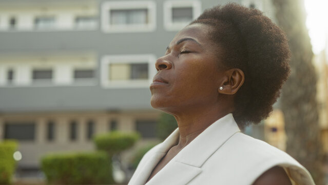 Woman with closed eyes tilting head upward, showing earring and neck, standing near modern building facade; calm reflection.