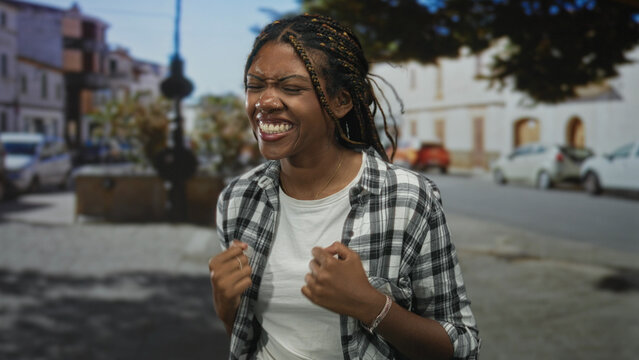 Woman with clenched fists pulling at her shirt while eyes closed and smiling on a sunny street; joy celebration.