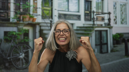 Naklejka premium Grey haired woman with bare shoulders and arms, hands together above head on a cobbled street in front of a building and bicycle; joyful celebration freedom.