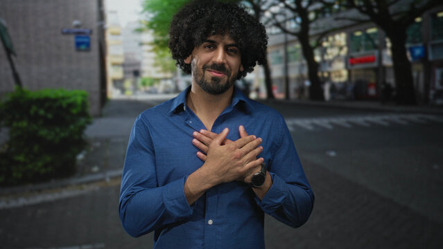 Man with hands crossed over chest on a city street wearing a blue shirt, curly hair and beard visible, smiling gently; gratitude sincerity calm.