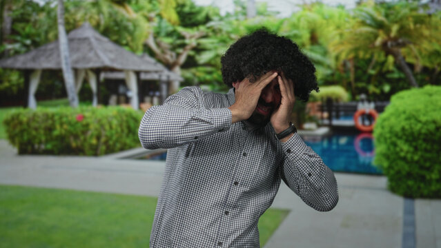 Man with curly hair pressing both hands to forehead at outdoor pool resort beside gazebo, bare hands covering eyes for headache; stress fatigue worry.