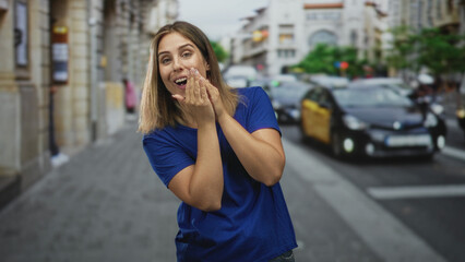 Fototapeta premium Woman with hands covering face on urban street, young blonde hispanic in blue shirt expresses covering gesture and laughter; surprise joy.