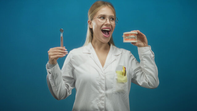 Woman dentist in white coat holding dental mirror and orthodontic braces model, mouth open showing teeth, studio portrait; cheerful confidence.