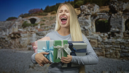 Naklejka premium Woman clutching multiple wrapped gift boxes, mouth open as she shouts while standing by ancient stone building ruins; joy celebration.