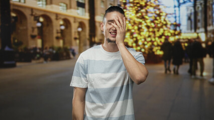 Fototapeta premium Man covering face with hand while smiling in a decorated building with blurred christmas tree and shoppers; embarrassed amusement.
