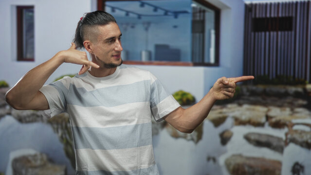 Man pointing finger upward with visible forearms in striped tshirt, slight smile, standing before a modern building courtyard; casual ease relaxed.