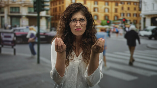 Woman with palms up and questioning hands gesture on a busy city street crosswalk, frowning and leaning forward while asking silently; confusion.