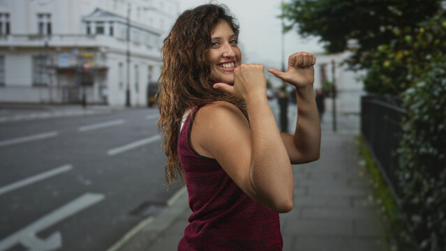 Woman with exposed shoulder and upper arm, thumbs pointing to chest while smiling on a city street with railing and pavement visible; confidence empowerment.