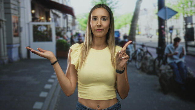 Woman with visible navel piercing and bare midriff, left palm up and right index finger pointing forward on a street with bicycles and shopfronts nearby; thoughtful.