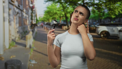 Fototapeta premium Woman holding makeup brush and touching neck with a pained squint on a city street; self care discomfort.
