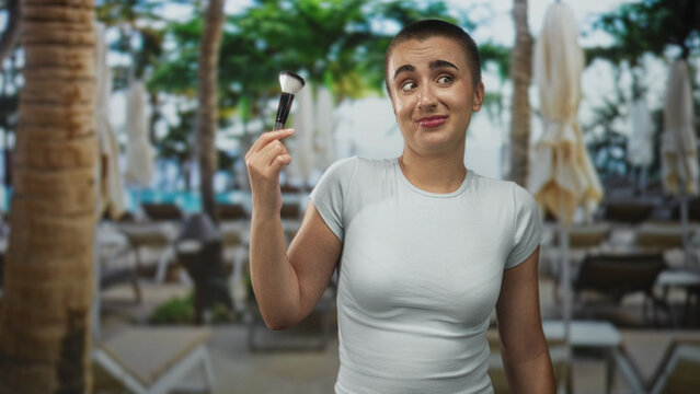 Woman with short hair holds makeup brush up near face with a half shrug expression in building; playful confusion.