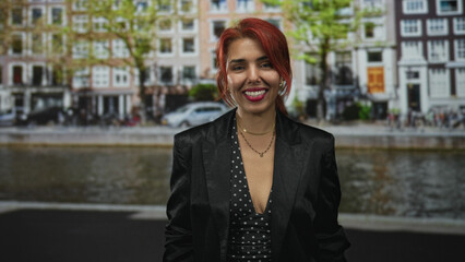 Fototapeta premium Woman smiling with visible cleavage, red hair and black blazer standing on canal street in amsterdam; confidence.